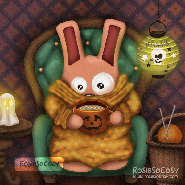 It’s an illustration of a pink Freezer Bunny with dark pink ears. Freezer Bunny is wearing an ochre yellow dotted snuggly blanket type sweater, holding an orange pumpkin mug with what looks like a latte. Freezer Bunny is seated on a teal coloured armchair with a diamond tuffed pattern on the back, and a wooden frame and legs. On the left is a round end table with a ghost candle holder. On the right is a wicker basket on the floor with little balls of yarn and knitting needles.