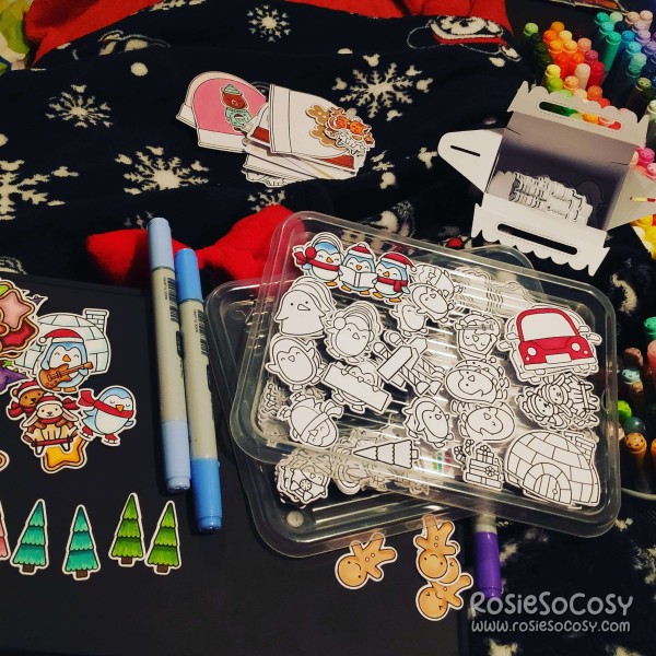 Rosie is sitting on the sofa, with craft supplies scattered everywhere. There are blankets on her legs, hundreds of stamped and cut images on lids, waiting to be coloured. Some images have already been coloured in with Copic markers. The images are all in a winter and Christmas theme; penguins, pine trees, Igloos, gingerbread cookies, snow globes.