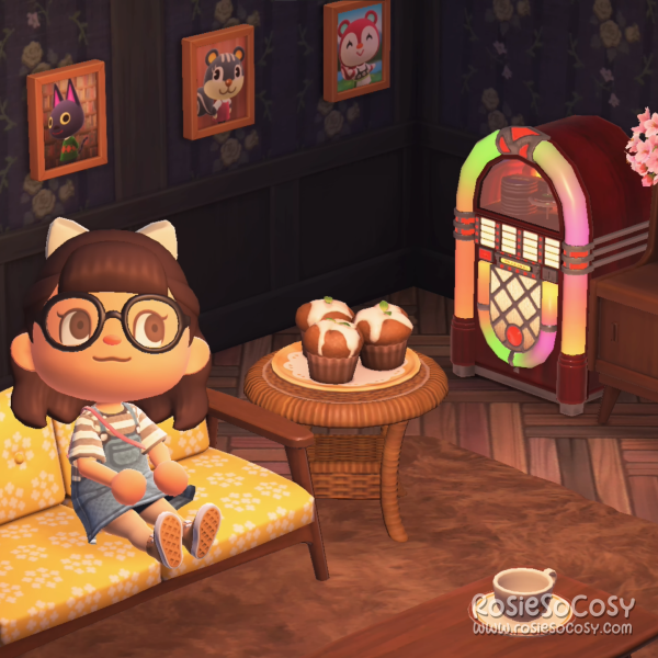Rosie in her living room in Animal Crossing: New Horizons. She's on a Nordic sofa with floral yellow cushions. Next to her is an end table with yummy cupcakes. The jukebox is playing Chillwave. All around her living room there are photographs of her villagers and NPCs. Rosie has dark brown hair, brown eyes and a white bow in her hair. She is wearing black rimmed glasses and a denim dress, with a grey and white striped shirt underneath.