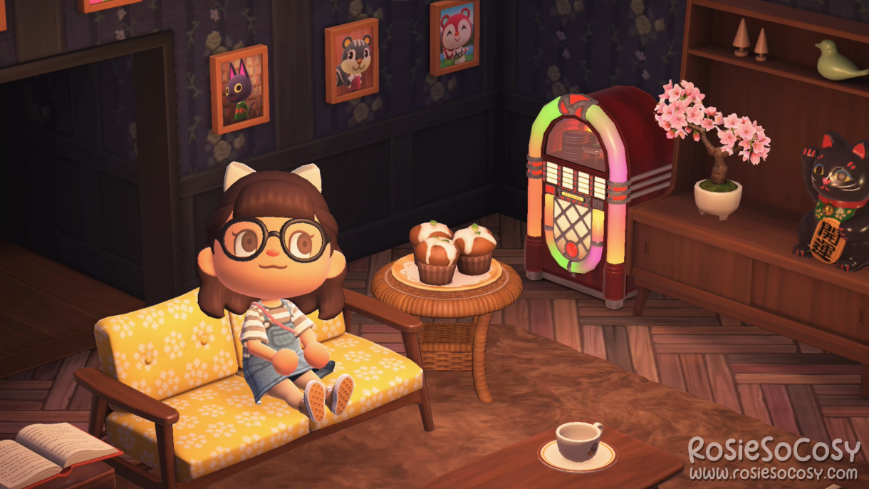 Rosie in her living room in Animal Crossing: New Horizons. She's on a Nordic sofa with floral yellow cushions. Next to her is an end table with yummy cupcakes. The jukebox is playing Chillwave. All around her living room there are photographs of her villagers and NPCs. Rosie has dark brown hair, brown eyes and a white bow in her hair. She is wearing black rimmed glasses and a denim dress, with a grey and white striped shirt underneath.