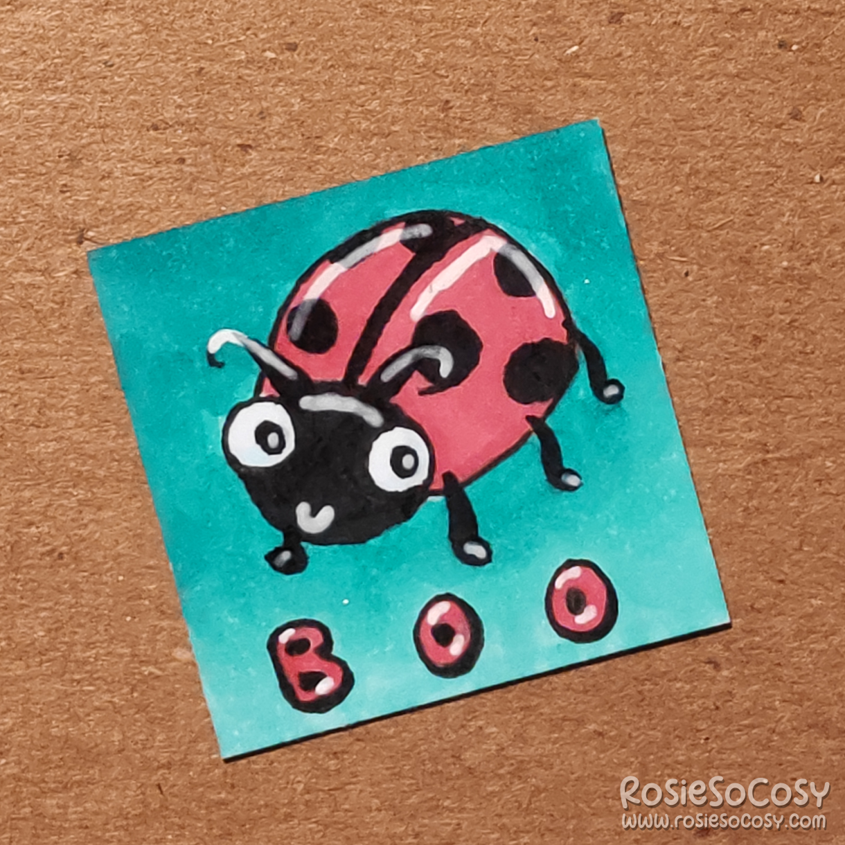 An inch sized drawing of a cute red and black ladybug. Underneath the drawing it says "boo" on a turquoise dark to light gradient background.