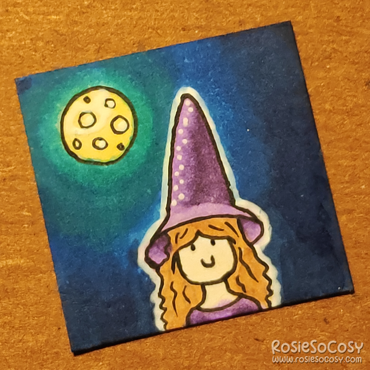 An inch sized drawing of a little witch/ She is wearing a purple outfit and pointy hat. It's dark outside and there is a full moon glowing in the background.