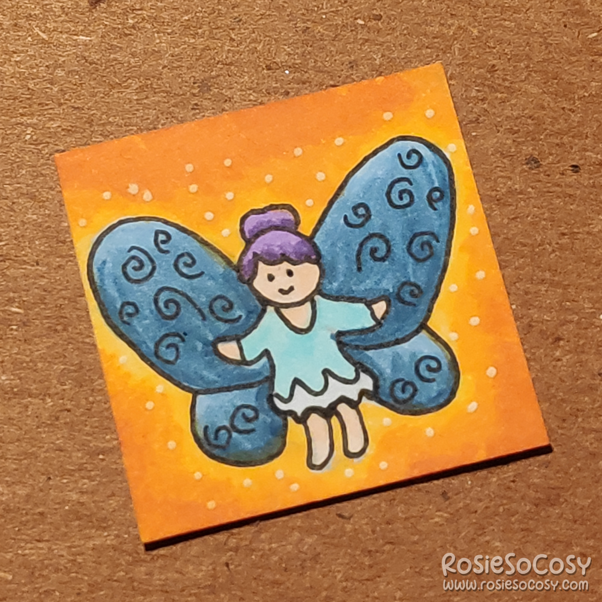 An inch sized drawing of a fairy with blue wings. She has a purple updo, a pastel blue tunic and a fair skintone. The background is orange and yellow with white glittery sparkles.