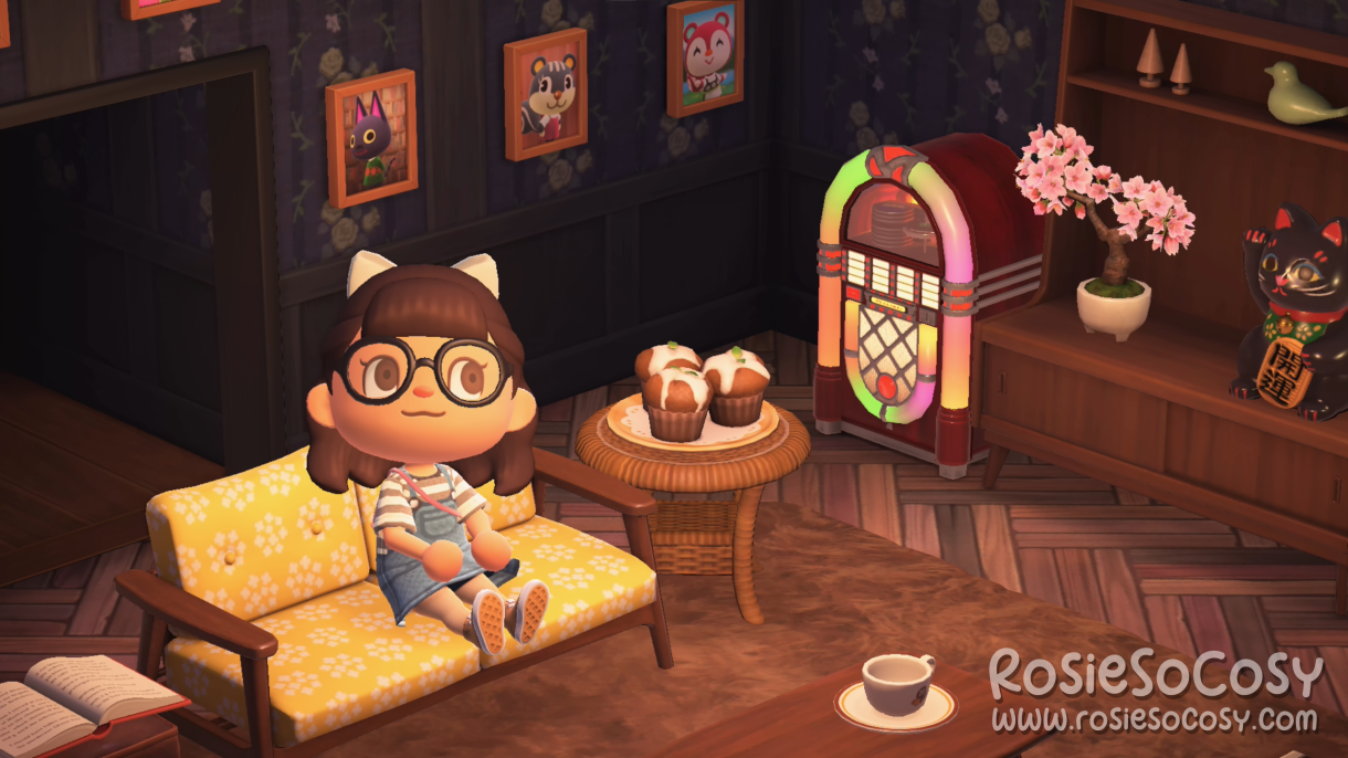 Rosie in her living room in Animal Crossing: New Horizons. She's on a Nordic sofa with floral yellow cushions. Next to her is an end table with yummy cupcakes. The jukebox is playing Chillwave. All around her living room there are photographs of her villagers and NPCs. Rosie has dark brown hair, brown eyes and a white bow in her hair. She is wearing black rimmed glasses and a denim dress, with a grey and white striped shirt underneath.