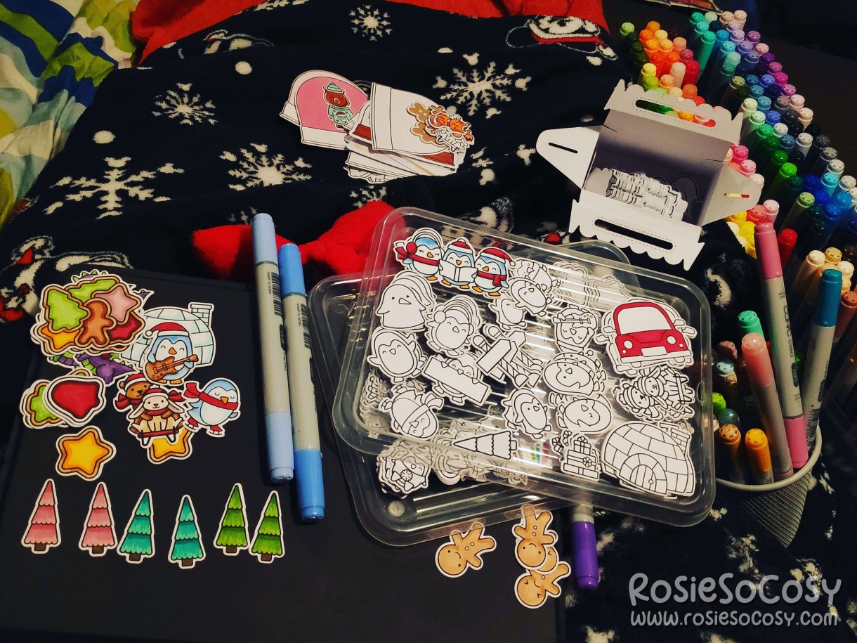 Rosie is sitting on the sofa, with craft supplies scattered everywhere. There are blankets on her legs, hundreds of stamped and cut images on lids, waiting to be coloured. Some images have already been coloured in with Copic markers. The images are all in a winter and Christmas theme; penguins, pine trees, Igloos, gingerbread cookies, snow globes.