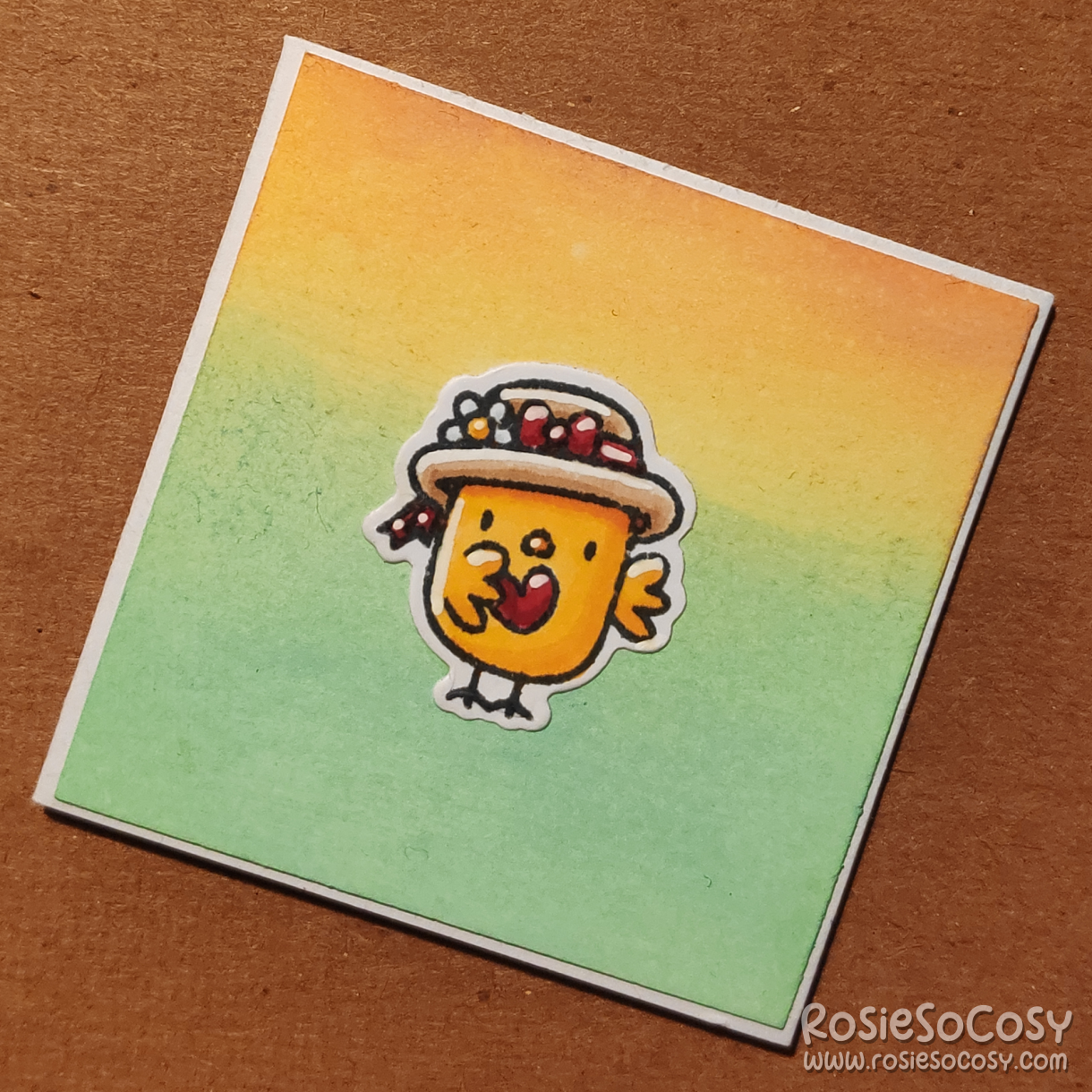 A inch sized card with a yellow to green gradient background. On the card is a tiny yellow chick with a cute floral hat, holding a tiny red heart.