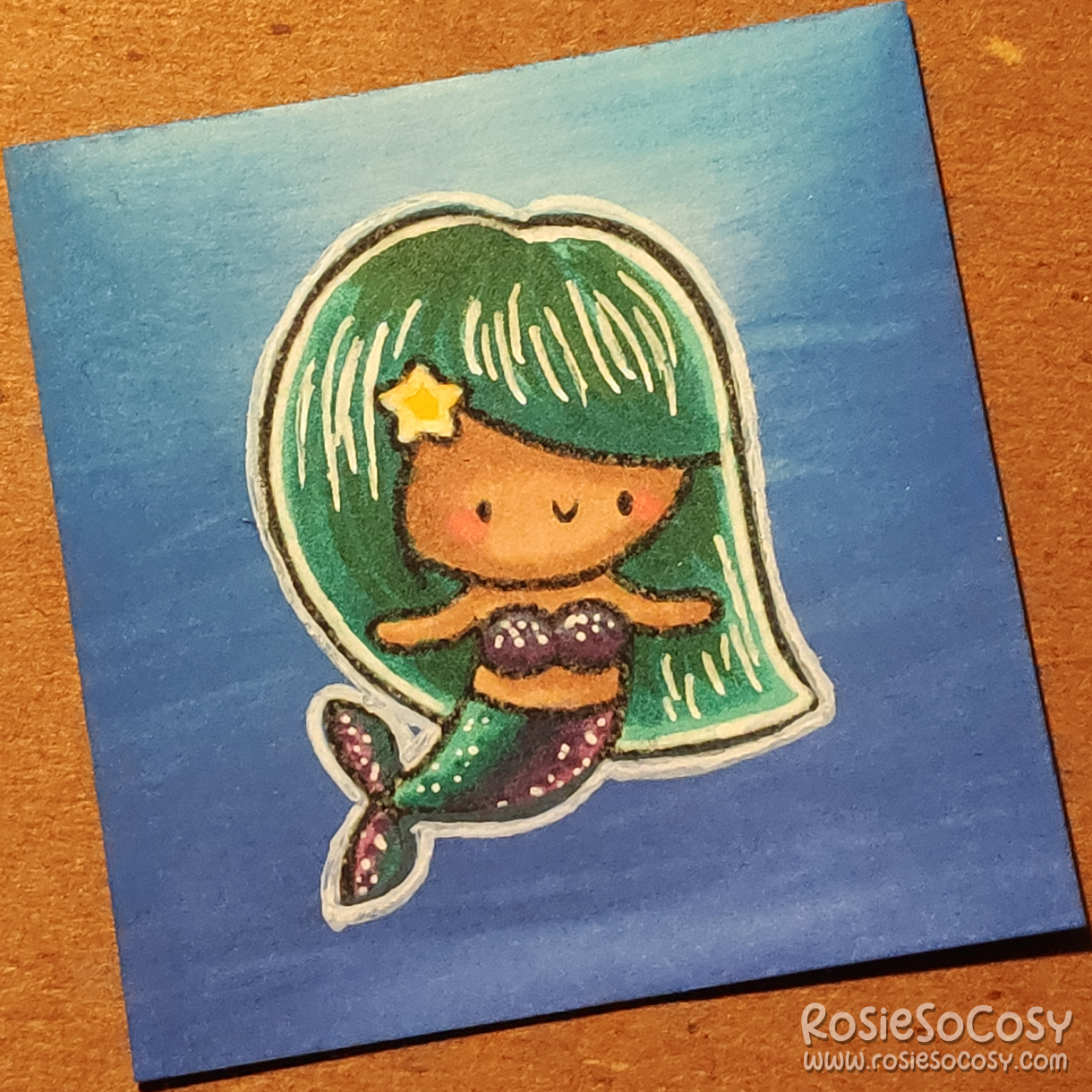 A tiny, 2 inch card with a cute mermaid on it. The mermaid is submerged in the blue sea, with light coming from above. The mermaid has seafoam/teal hair, and a purple/teal tail/top. She has a medium/tan skin tone and a cute yellow star in her hair.