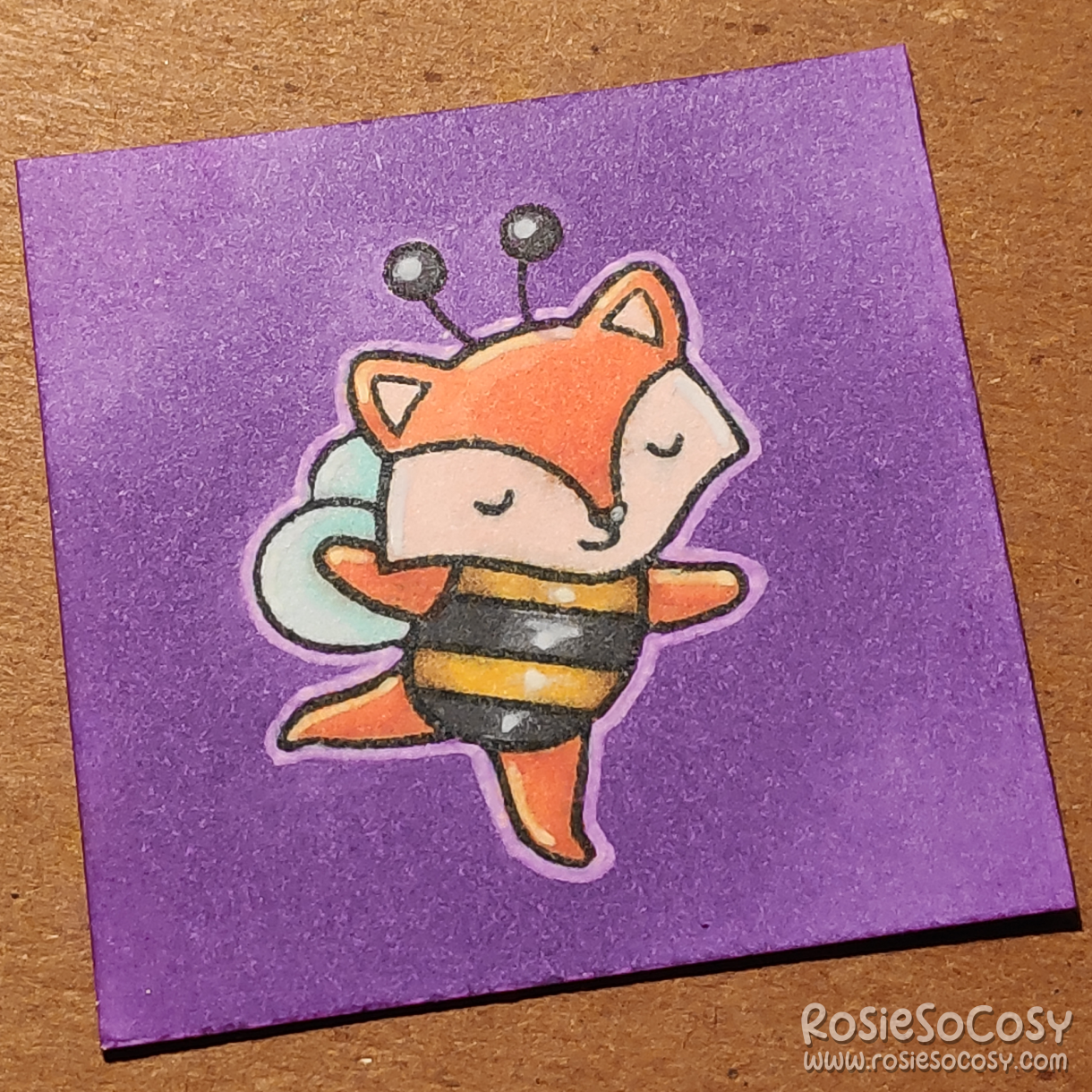 A tiny 2 inch card with an orange fox dressed up in a black and yellow bee costume. The fox is dancing with their eyes closed. The background is purple.