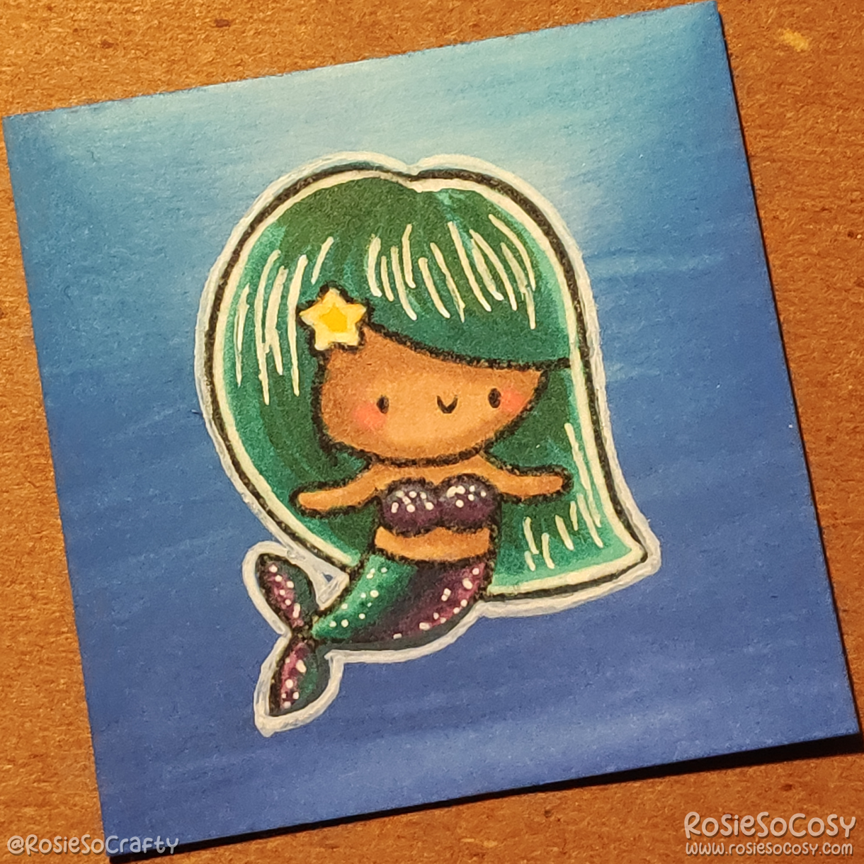 A tiny, 2 inch card with a cute mermaid on it. The mermaid is submerged in the blue sea, with light coming from above. The mermaid has seafoam/teal hair, and a purple/teal tail/top. She has a medium/tan skin tone and a cute yellow star in her hair.