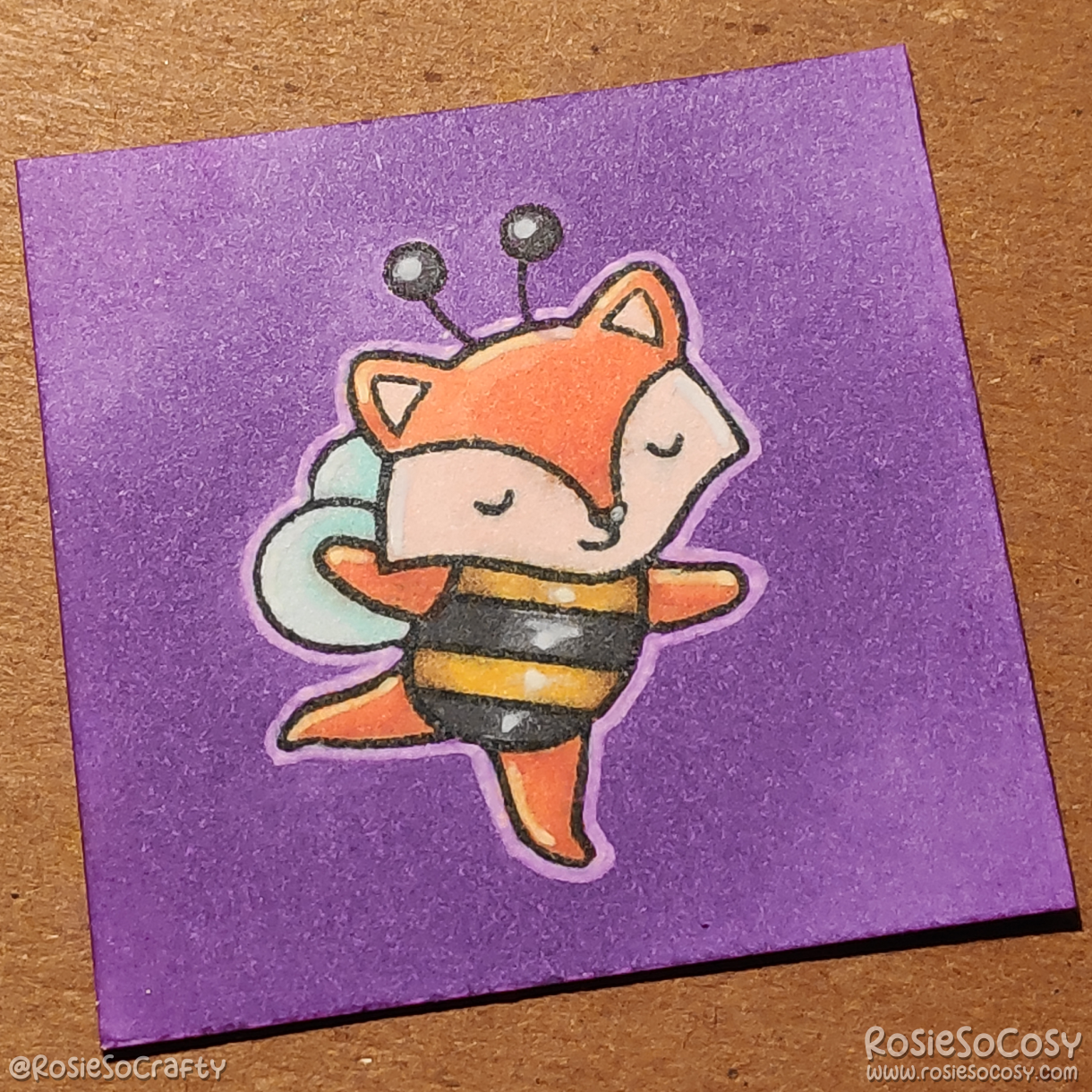A tiny 2 inch card with an orange fox dressed up in a black and yellow bee costume. The fox is dancing with their eyes closed. The background is purple.